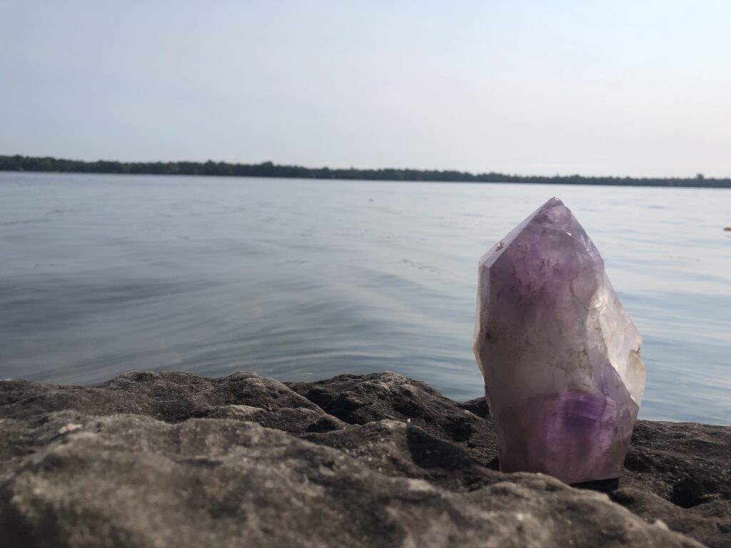 Close up of large amethyst crystal point standing on rocks next to a calm lake